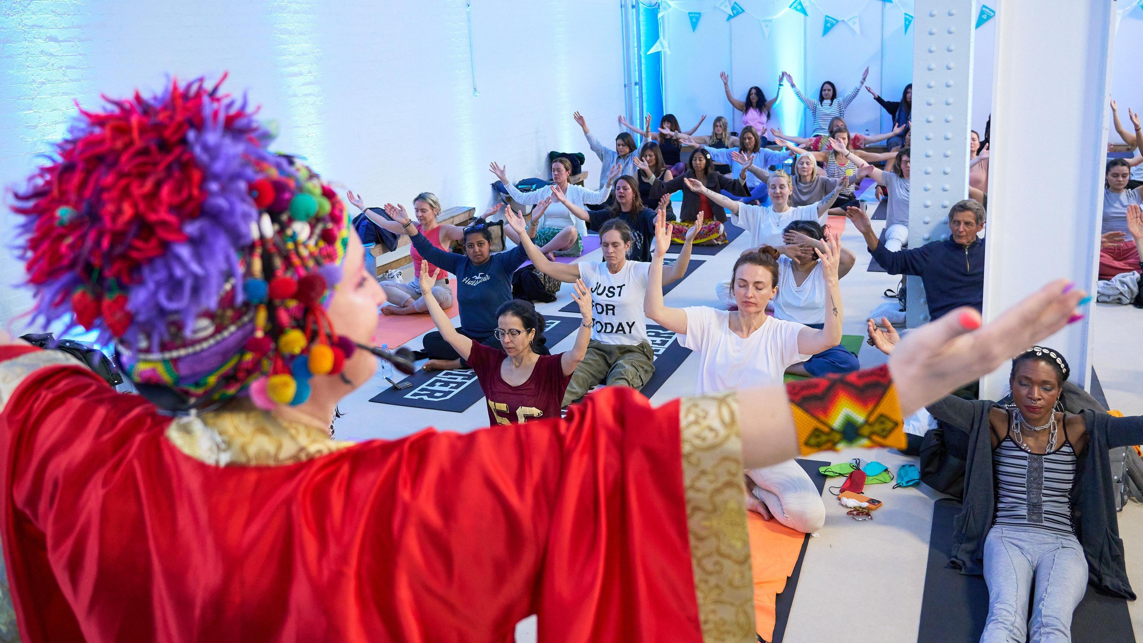 A woman is wearing a red and purple coloured hat along with a red shawl. She has her arms pointed outwards to the sky. People are sitting on the floor on yoga mats, copying her arm actions.