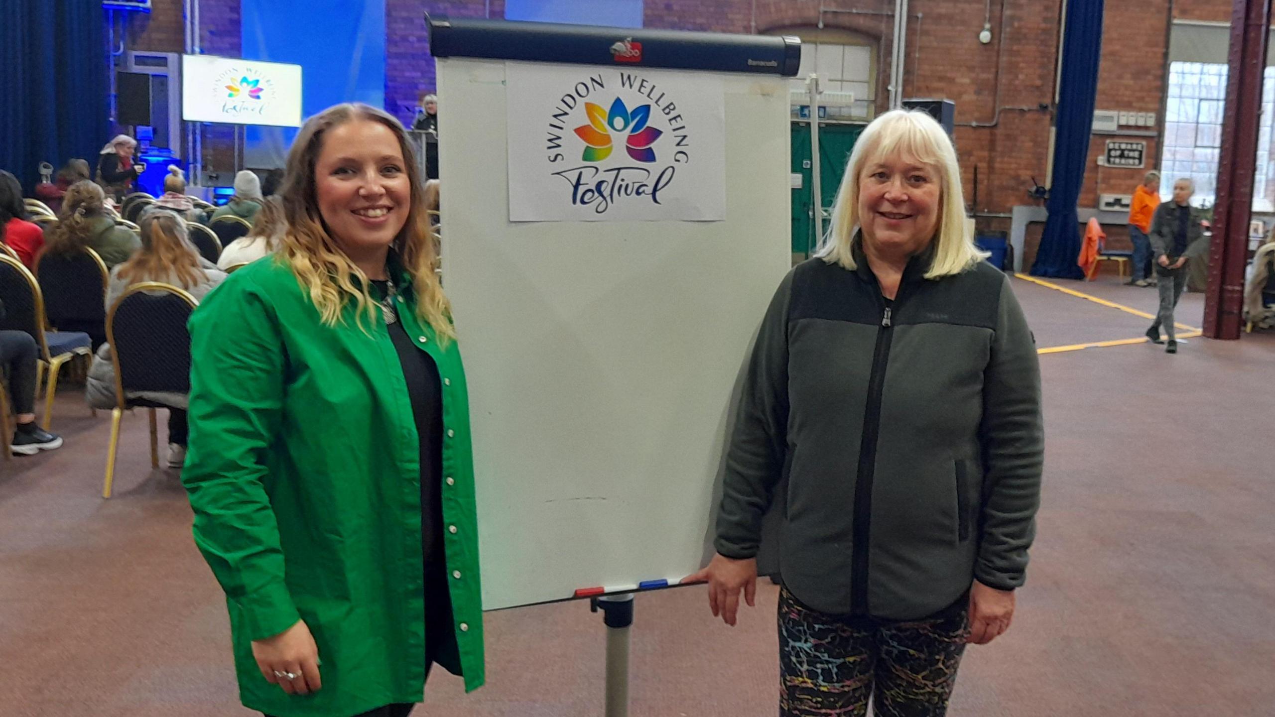 Sian Baxter, an alignment and breathwork coach, stands on the left, wearing a green shirt. Event organiser Sally Langheim is wearing a dark jacket and the two women are smiling at the camera next to a Swindon Festival A-board. There are people in the background sitting watching a presentation.