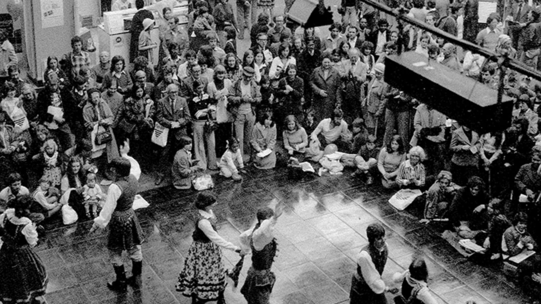 Crowds sit around a stone dance floor as people dressed in long white shirts and skirts dance together.