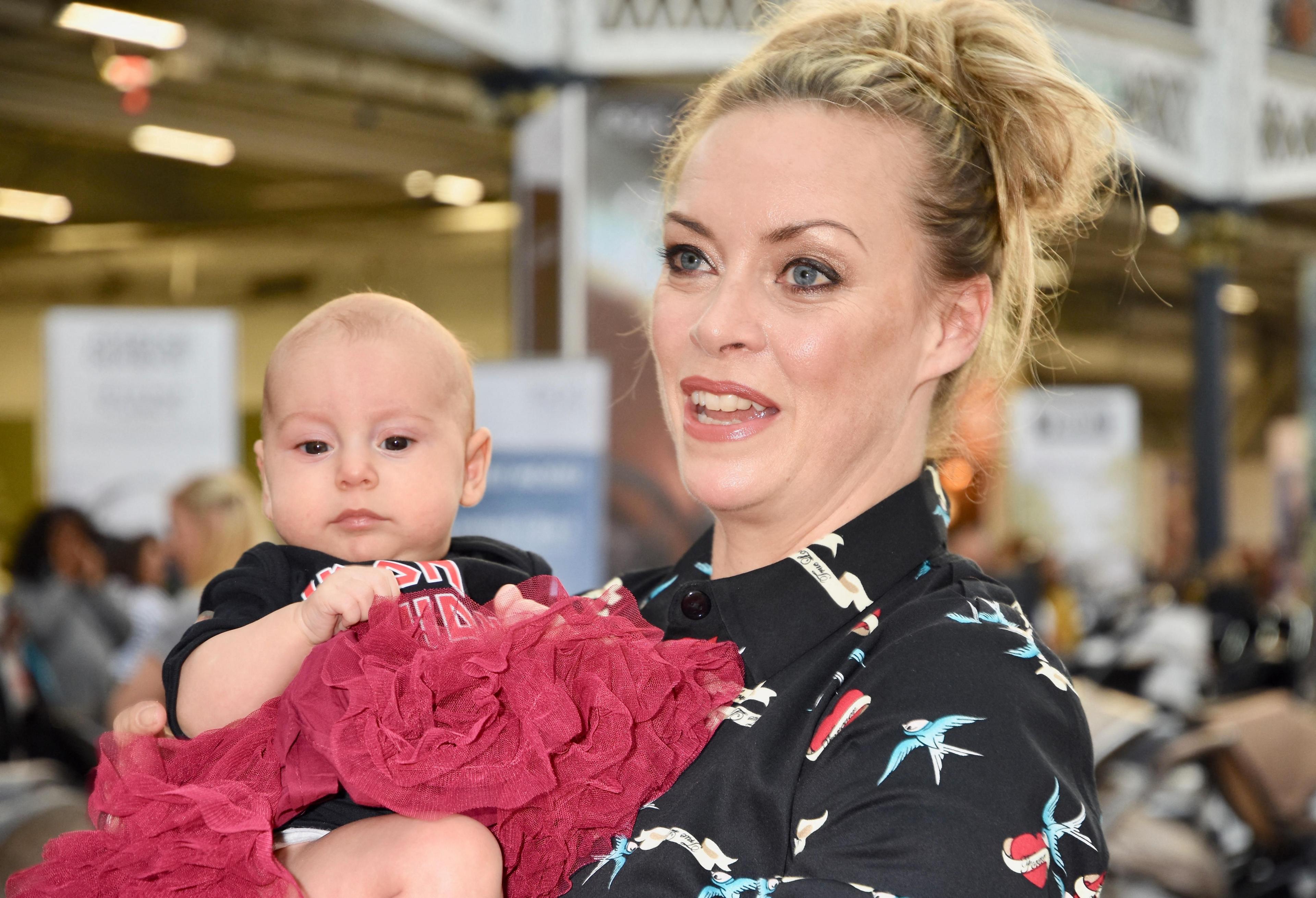 A woman with blonde hair loosely tied in a bun, and wearing a black shirt with birds and hearts printed on it, holds a baby, wearing a black tee and red tutu, close to her chest.