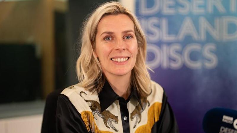 Sara Pascoe, who has shoulder-length blonde hair and wears a fringed black shirt with leopards on it, smiles as she sits in the Desert Island Discs studio
