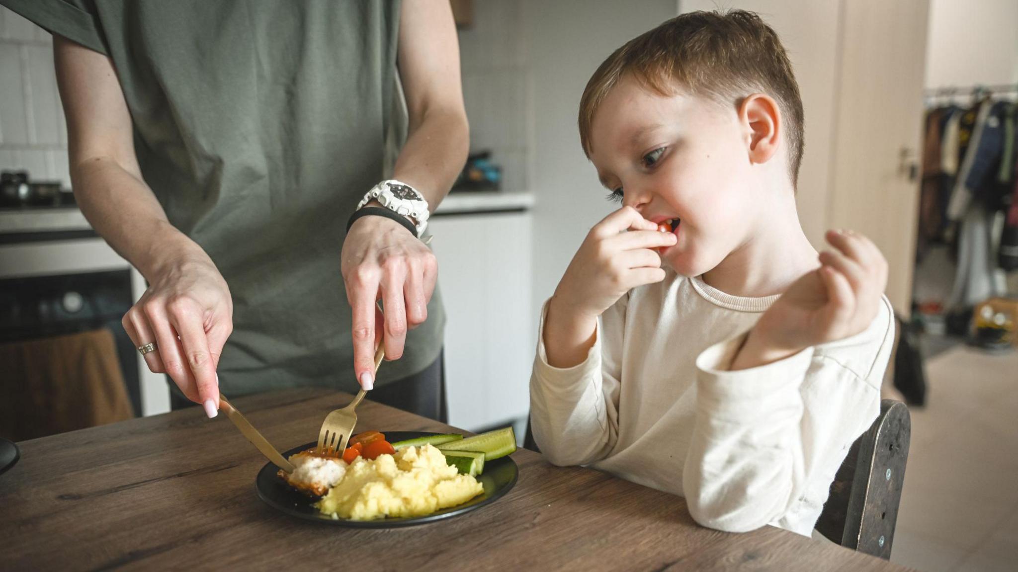 A young child with brown hair and a white top, eating a tomato, with a woman wearing a green top cutting up a plate of food in front of him.