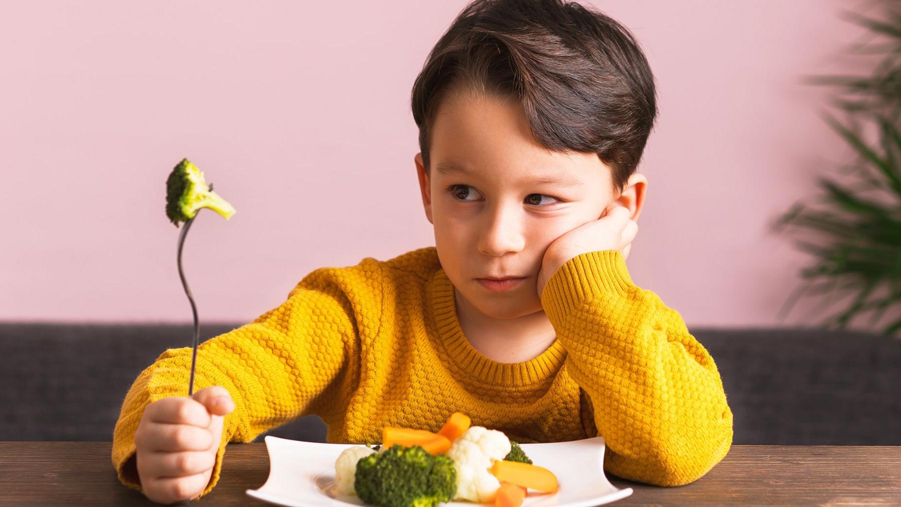 Young boy in yellow jumper sits at a table with a plate of broccoli, carrots and cauliflower in front of him. He is holding a fork with broccoli on it and looks unhappy.