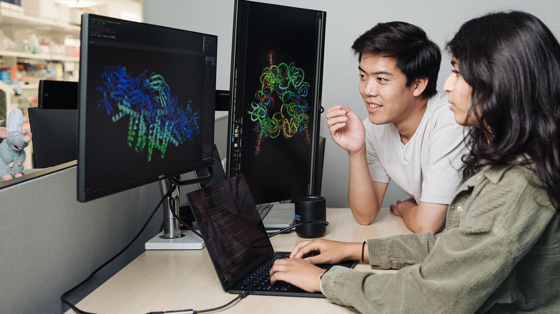 Two people working together at a desk in what appears to be a laboratory or research office environment. Large computer monitors display colorful three-dimensional molecular or protein structure models against dark backgrounds, while a laptop on the desk shows lines of code or data. One person is seated using the laptop, and another is leaning toward the screens as both focus on the scientific visualizations. Laboratory shelving and equipment are visible in the background, suggesting a biomedical, biotechnology, or computational research setting. The scene emphasizes the use of computer-based analysis and visualization tools to study complex biological structures.