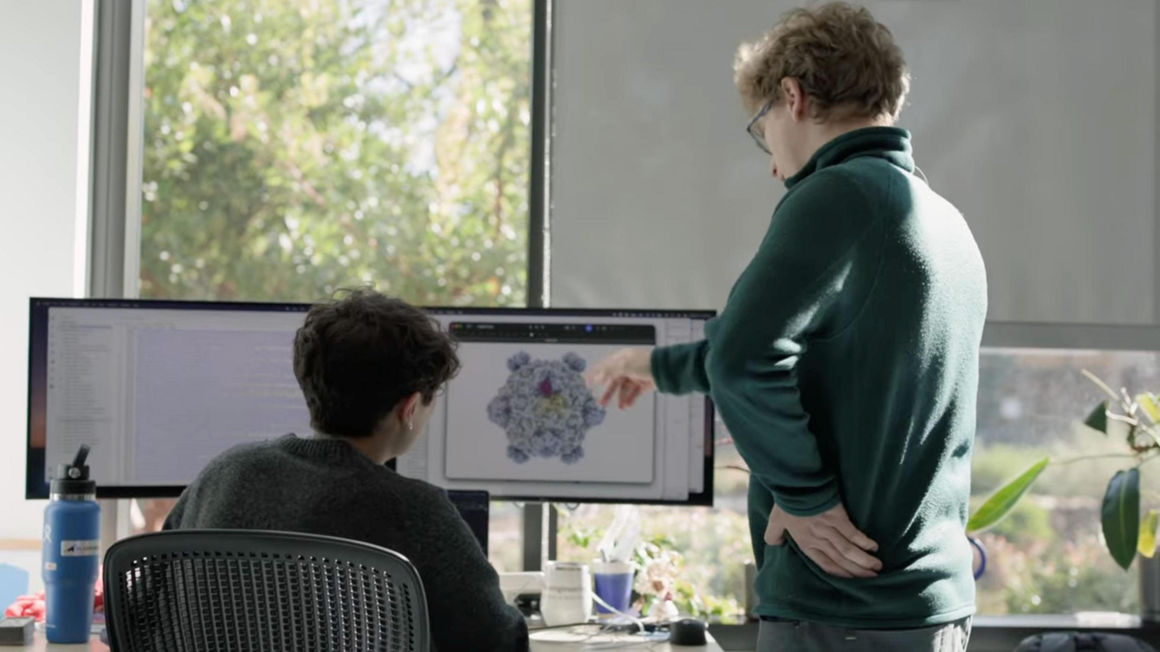This image shows two people working at a desk in a bright office or research environment with large windows and natural light. One person is seated in front of a dual-monitor workstation, while another stands nearby pointing toward a detailed molecular or protein structure displayed on one of the screens. The monitors show scientific visualization software, with one screen displaying a complex three-dimensional biological model and the other containing text or data. Various desk items, including a water bottle, mug, computer peripherals, and potted plants, are visible around the workspace. 