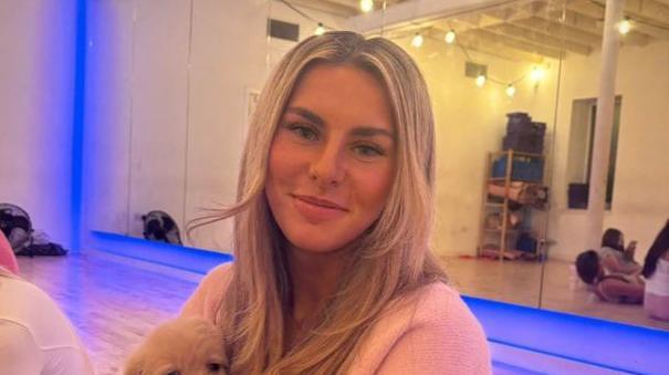 Ami Robertson sits in a Pilates studio. She is holding a dog on her lap and is smiling at the camera. She has long blonde hair.