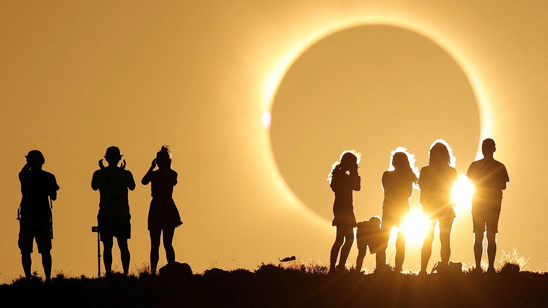 A total solar eclipse is about to occur - a darkened grey sphere appears large in the sky and bright yellow light is emanating from it drenching the sky. Two groups are gathered watching it, silhouetted against the sky