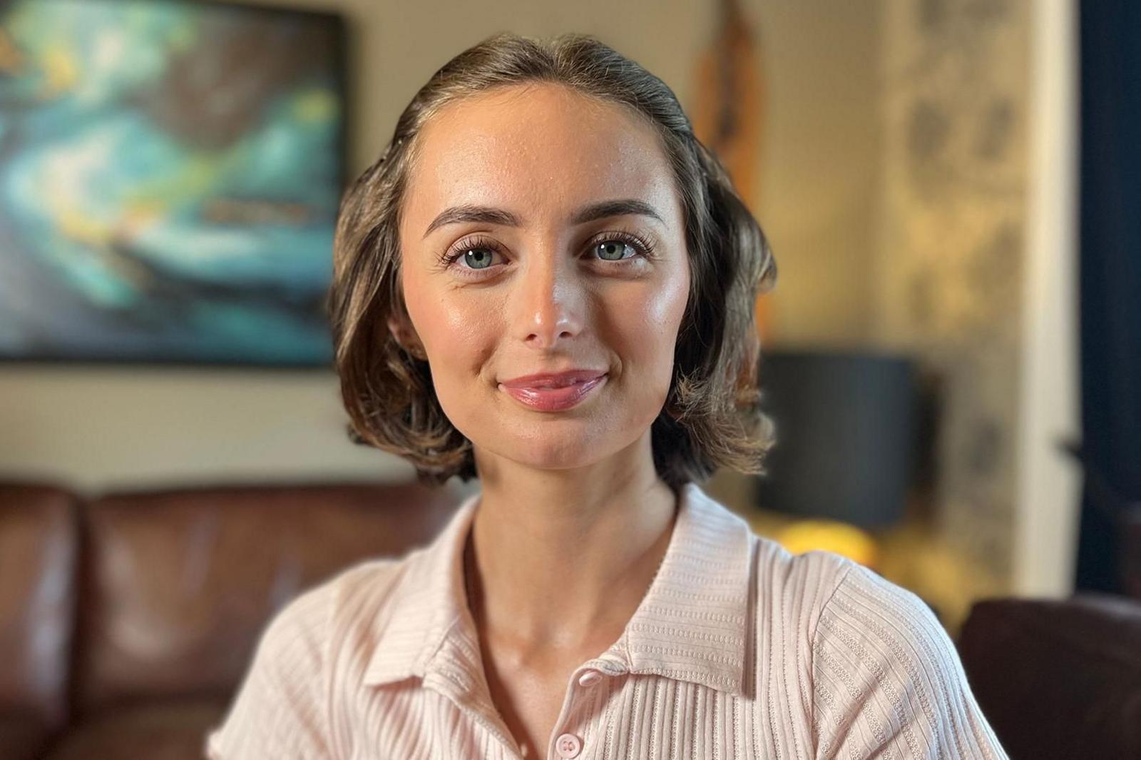 A woman seated indoors on a brown leather sofa, wearing a light pink ribbed collared top with a clip-on microphone attached. A framed abstract painting hangs on the wall in the blurred background, with soft indoor lighting and shallow depth of field creating a professional interview-style setting.