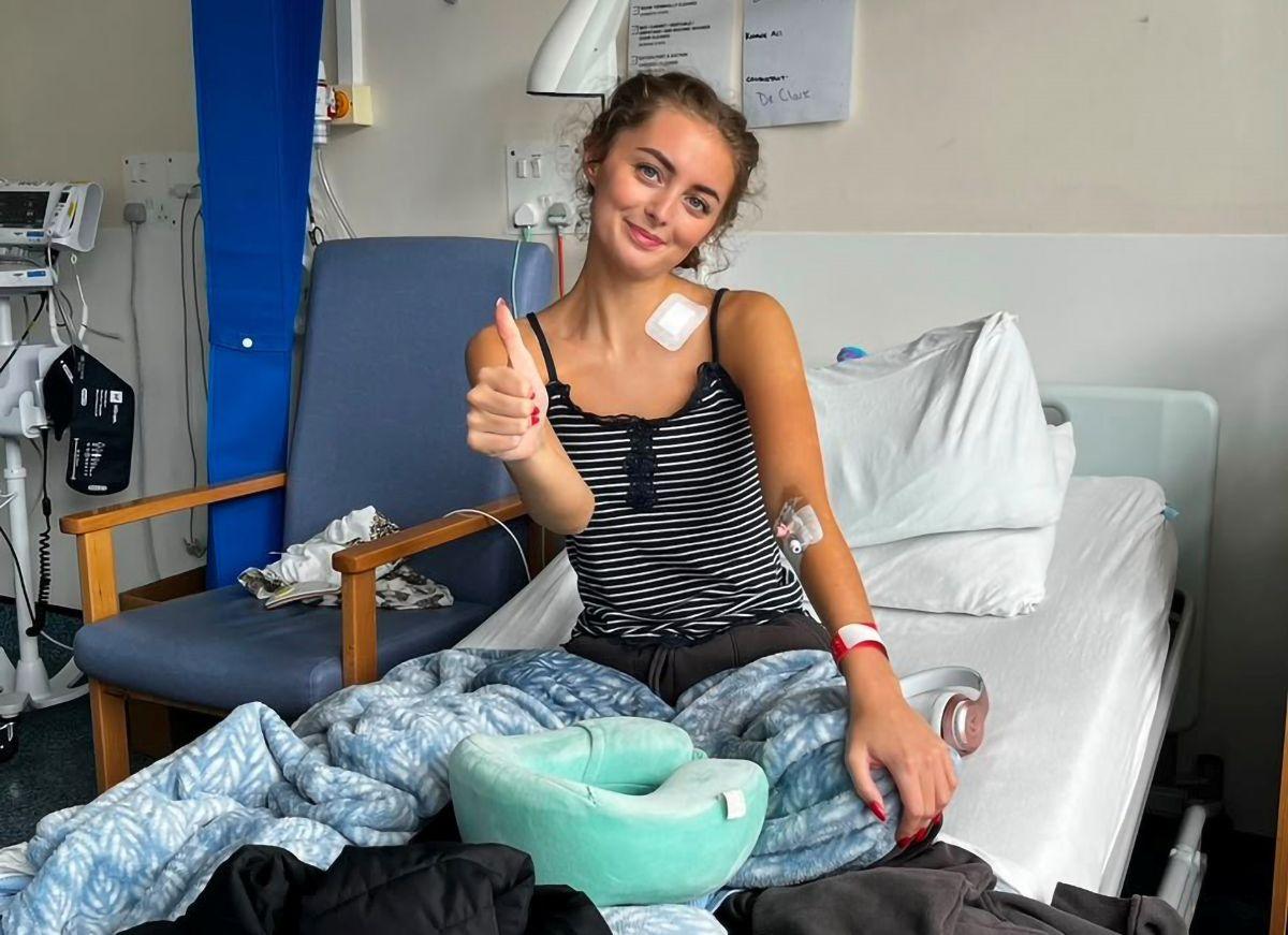 Person sitting upright in a hospital bed with a blanket over their legs, giving a thumbs-up gesture. Medical monitoring equipment, a chair, and hospital room furnishings are visible in the background. A travel neck pillow rests on the bed in the foreground.
