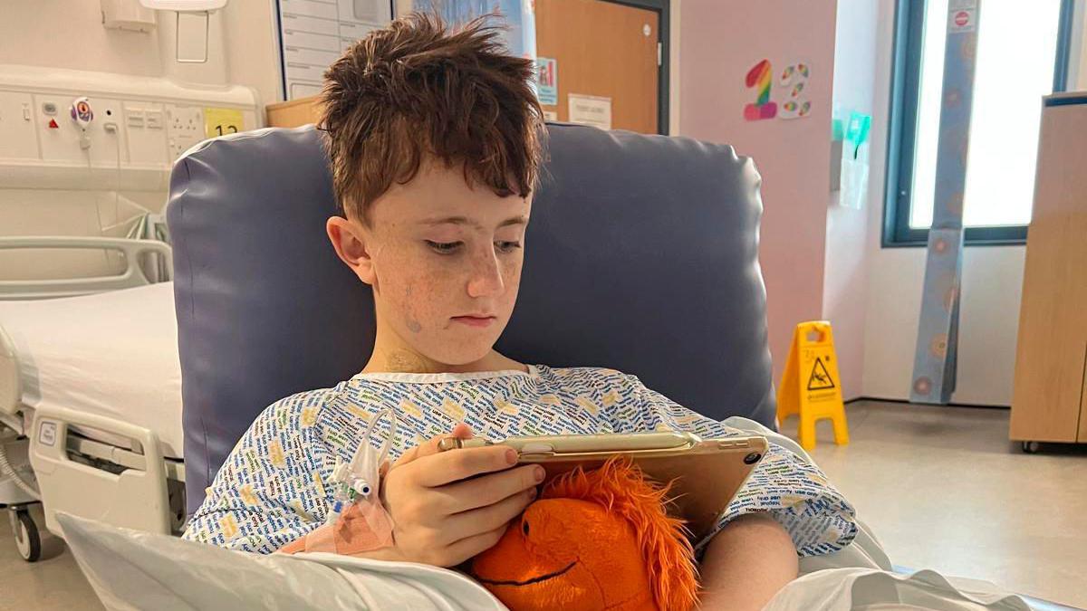 A young boy sitting his a chair wearing a hospital gown, he is looking down at an ipad and holding an orange soft toy,