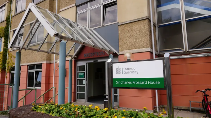 Outside of the States of Guernsey's Sir Charles Frossard House. The concrete building, at least three storeys tall, has a white and green sign outside. The ground floor has terracotta-coloured cladding.