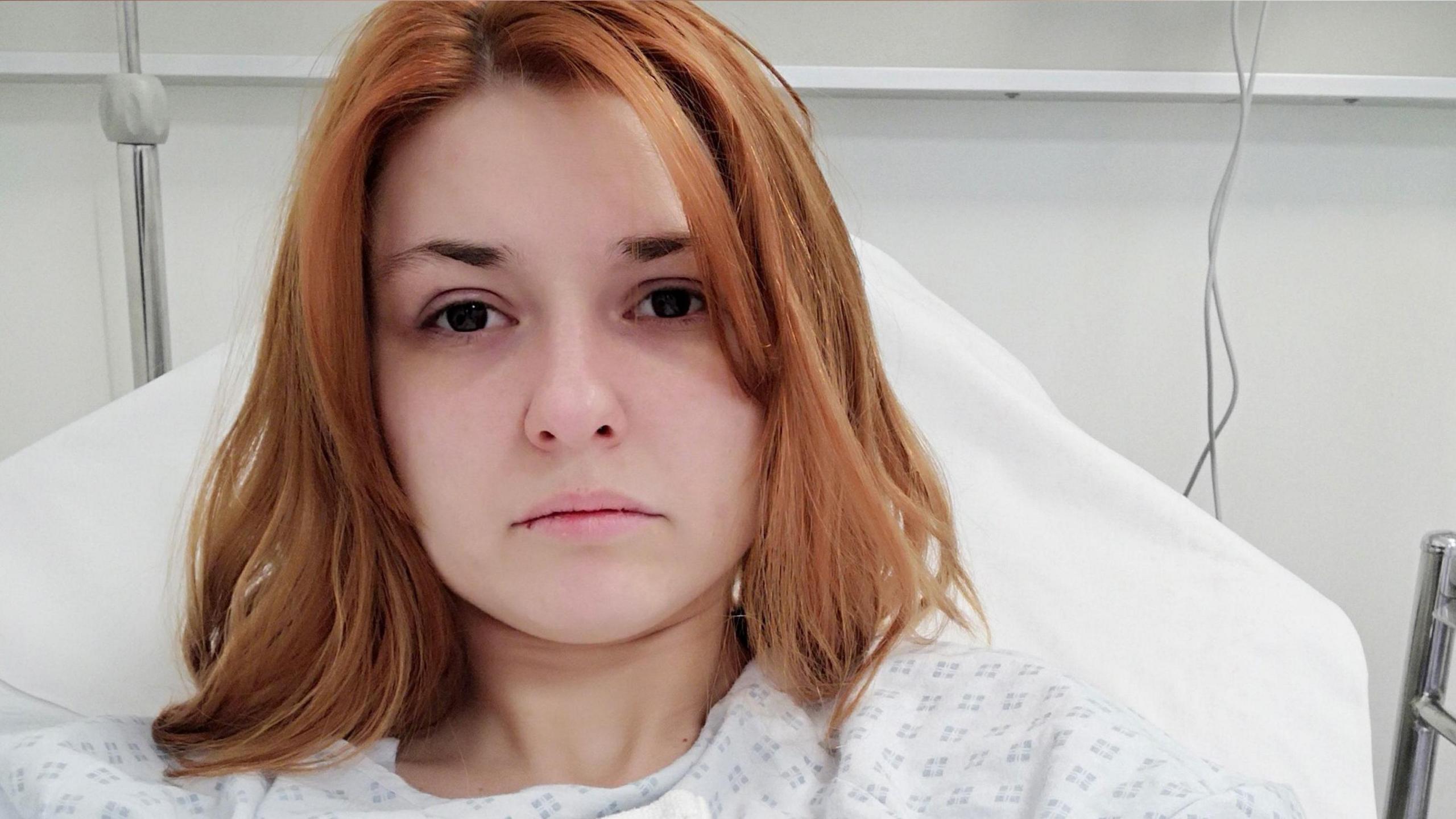 A woman sat in a hospital bed staring at the camera, she has ginger hair.