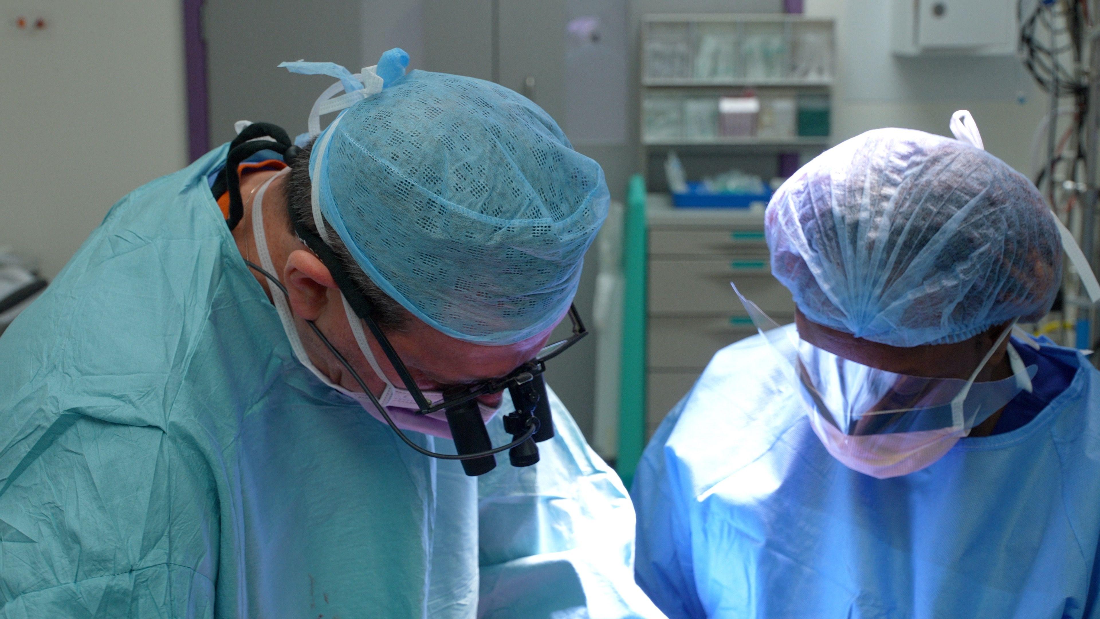 Two members of the surgical team at the Royal Papworth Hospital perform a double lung transplantation. The clinicians, a man and a woman, are wearing mint green and blue scrubs and hair nets. In the background we see elements of the operating theatre out of focus
