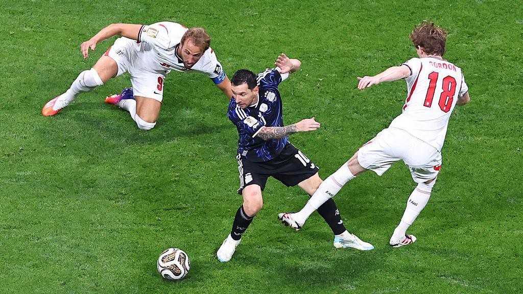 Lionel Messi of Argentina in action with Harry Kane and Anthony Gordon of England during Argentina's World Cup semi-final win against England. Kane is on the ground and Gordon's left leg looks to be swinging towards Messi as the England players, in white, try to dispossess Argentina's captain, in dark blue.