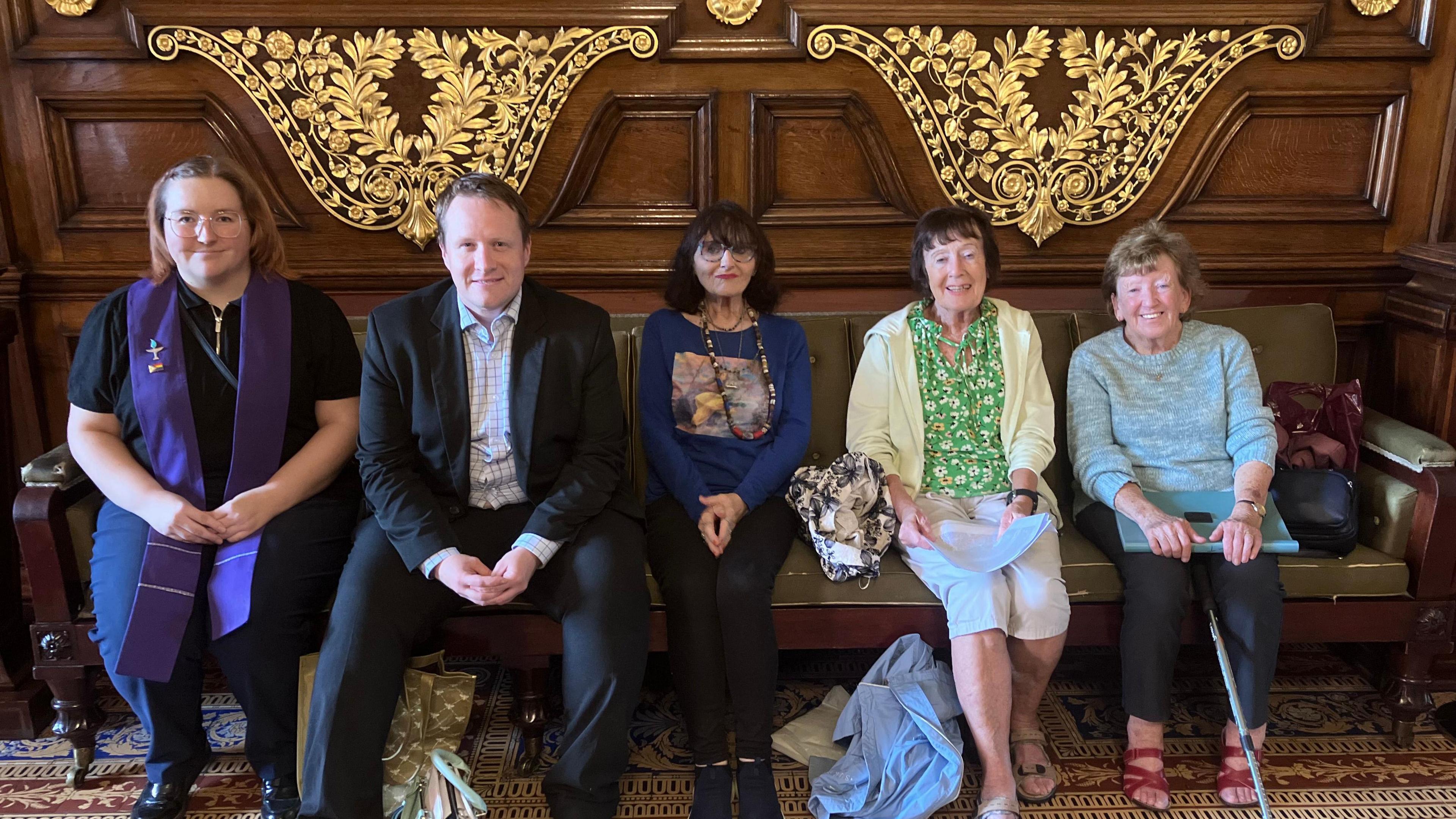Kris Brown sits inside with Gina and others, smiling to the camera.