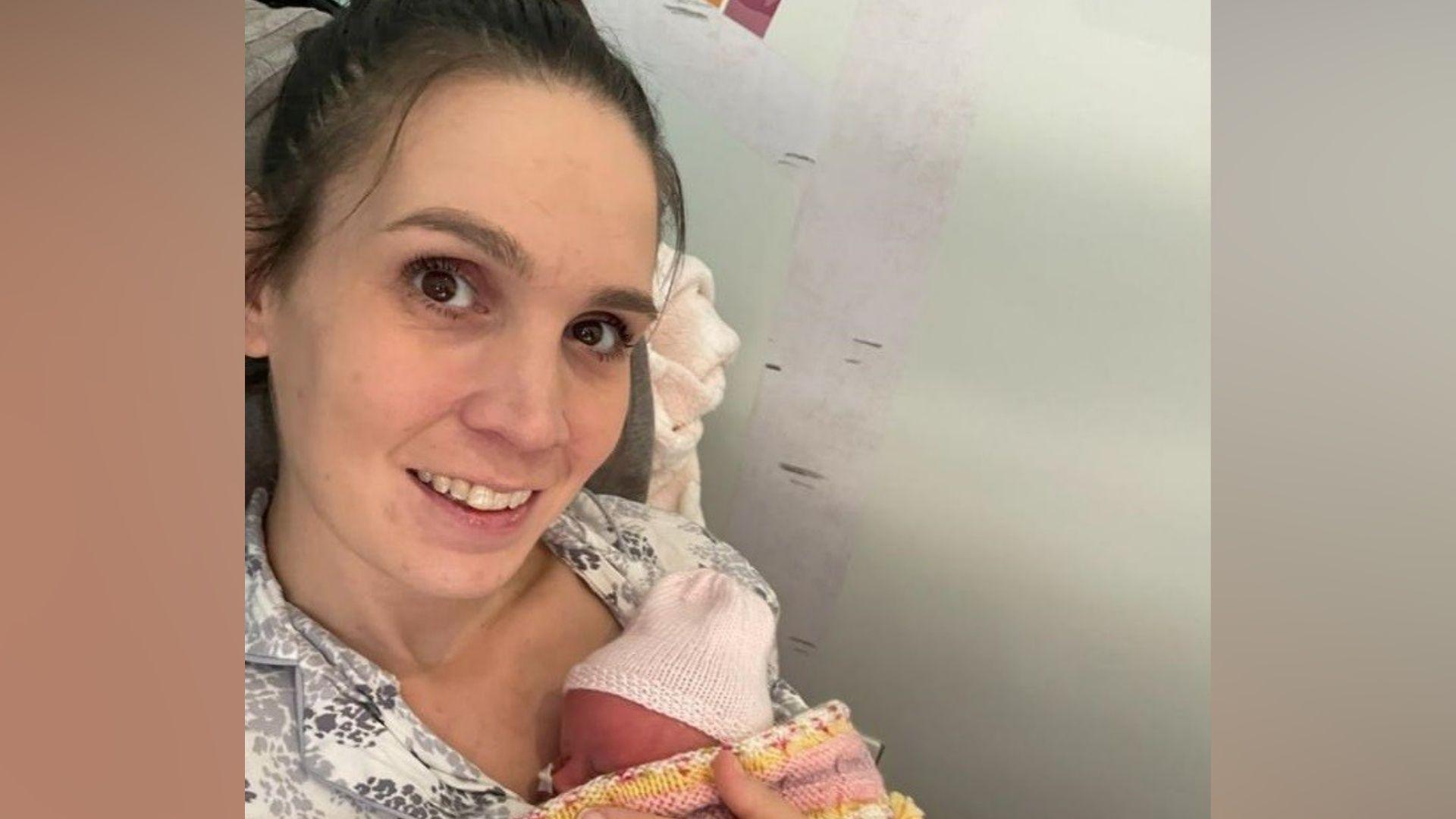Natalie is seated indoors holding a newborn baby wrapped in a pink, yellow and white knitted blanket. The baby is wearing a light-coloured knitted hat and is nestled against the person's chest.