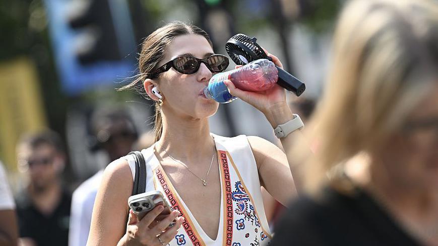 A woman wearing a sleeveless top and sunglasses holds a battery-operated fan in her hand while she drinks from a bottle of water. 