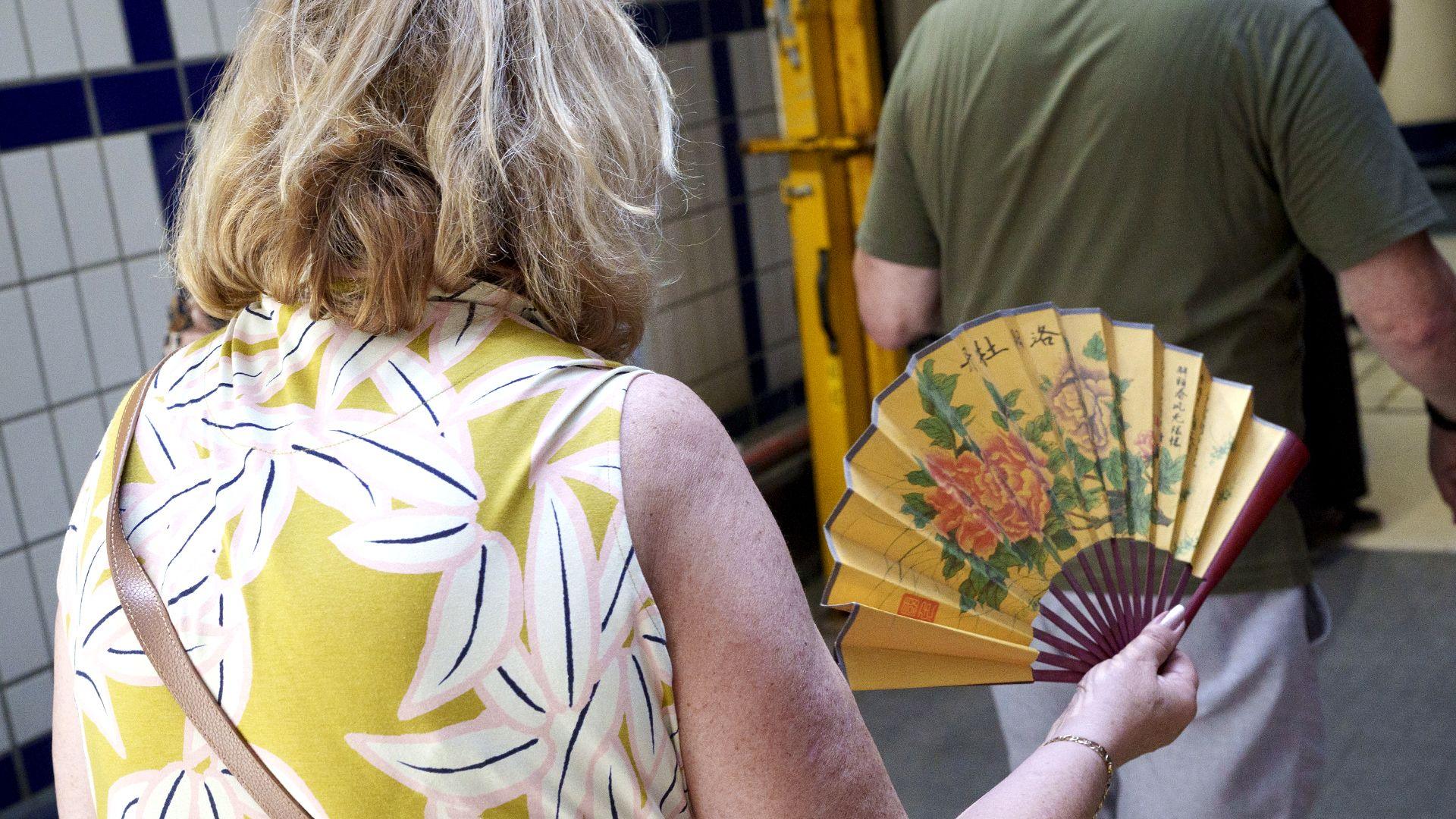 A lady in a white and yellow patterned dress enters a London Underground station with an oriental fan in her hand