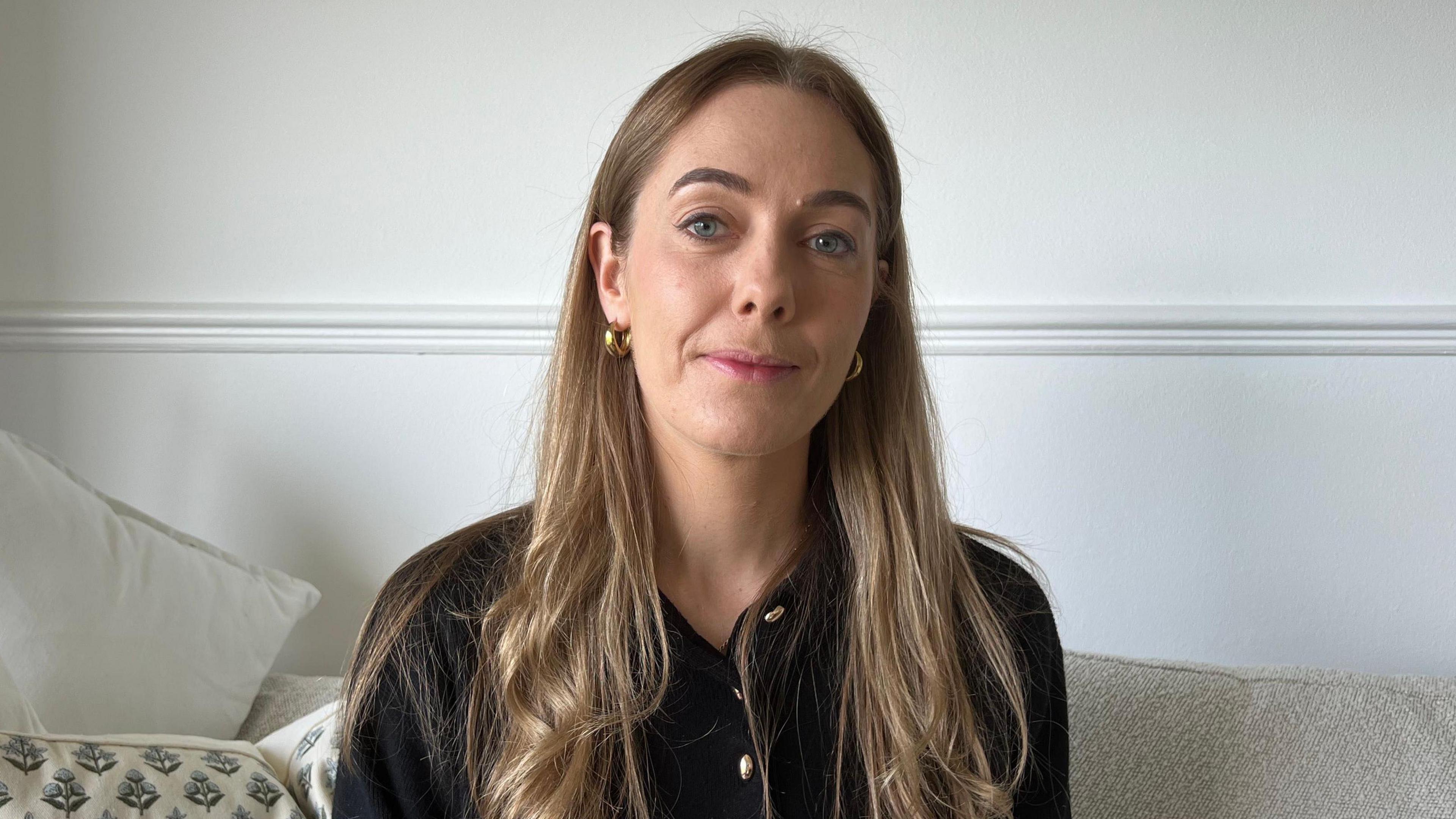 Jessica Pinel is sitting on a sofa, smiling at the camera. She has long blonde hair and is wearing a black blouse with gold buttons.