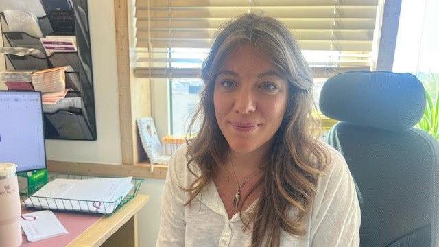 Dr Jessica Langtree-Marsh is sat in her clinic. She is wearing a white shirt and has brown hair. She is sat next to a computer.