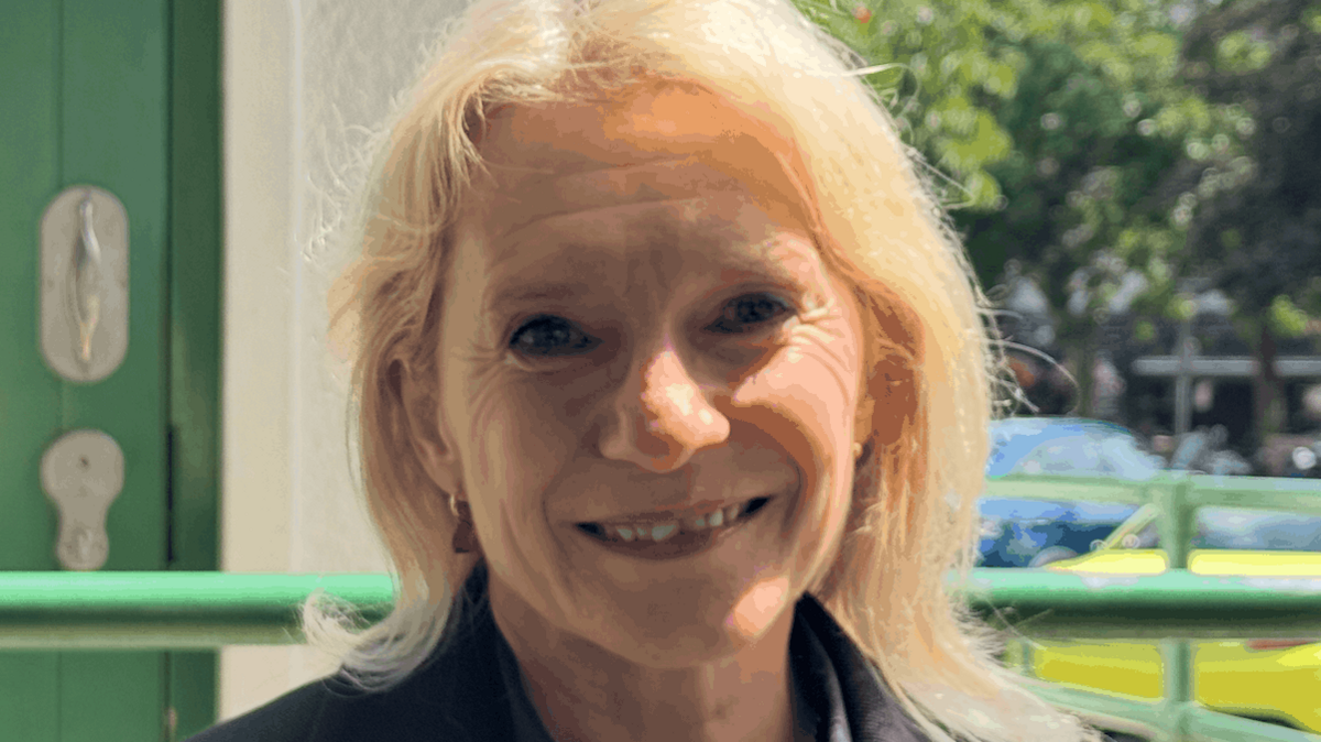 Senator Elaine Millar wearing a dark jacket, standing outside a public toilet entrance, with a green accessible restroom door visible in the background.