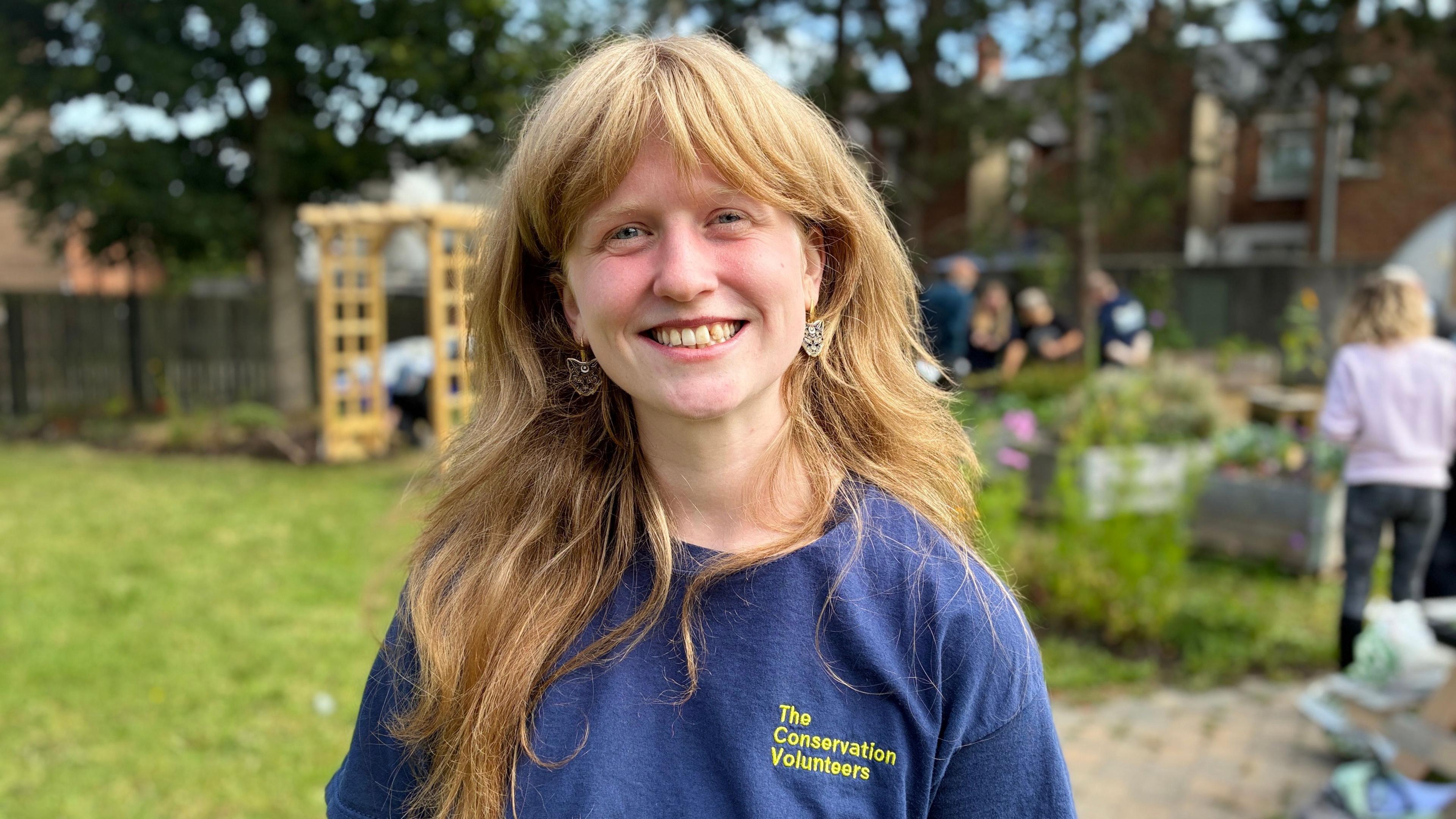 A young woman with long, light brown hair smiles brightly in a community garden. She’s wearing a navy blue T-shirt with yellow text reading “The Conservation Volunteers.” Behind her, other people are tending to plants and chatting near raised beds and a wooden garden arch. The scene is lively and green, reflecting teamwork and outdoor engagement.