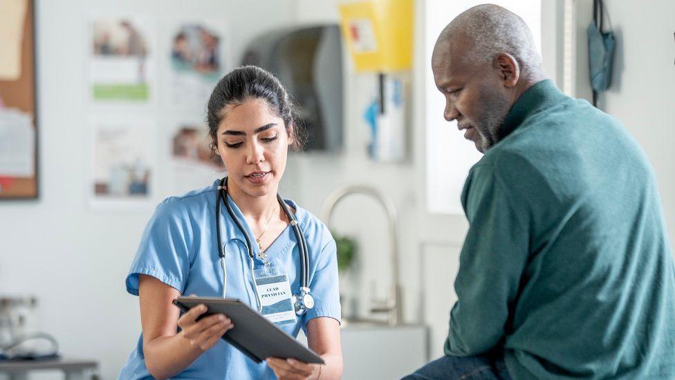 A nurse in blue scrubs with a stethoscope consults with a Black patient in a clinic, using a tablet. Warm, professional healthcare scene highlighting patient care and medical teamwork.