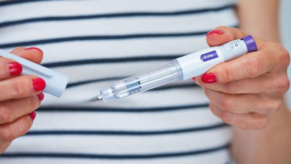 A woman with red nail polish wearing a stripey top holds a weight loss syringe.