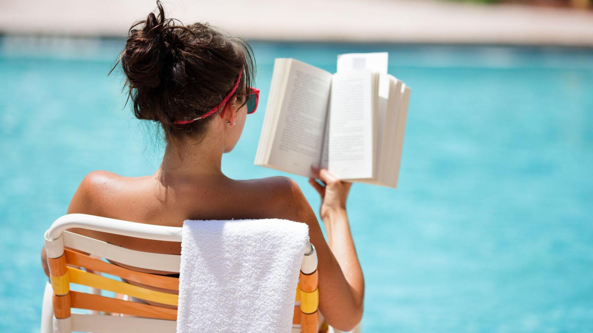 Young woman sitting in a chair with a towel draped over the back, reading at the pool in the summer