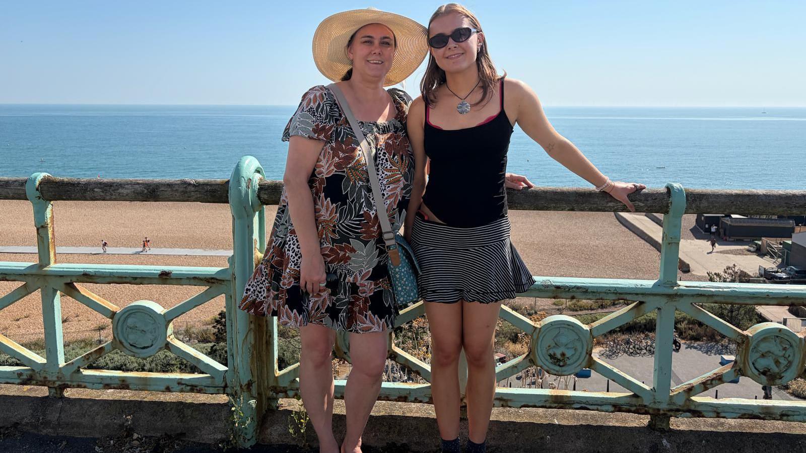 Caroline (L) stood next to her daughter (R). They are leaning on an ornate railing with their backs to a beach looking out to the sea. Caroline is wearing a dress and large summer hat and her daughter is wearing a black top and a skirt.