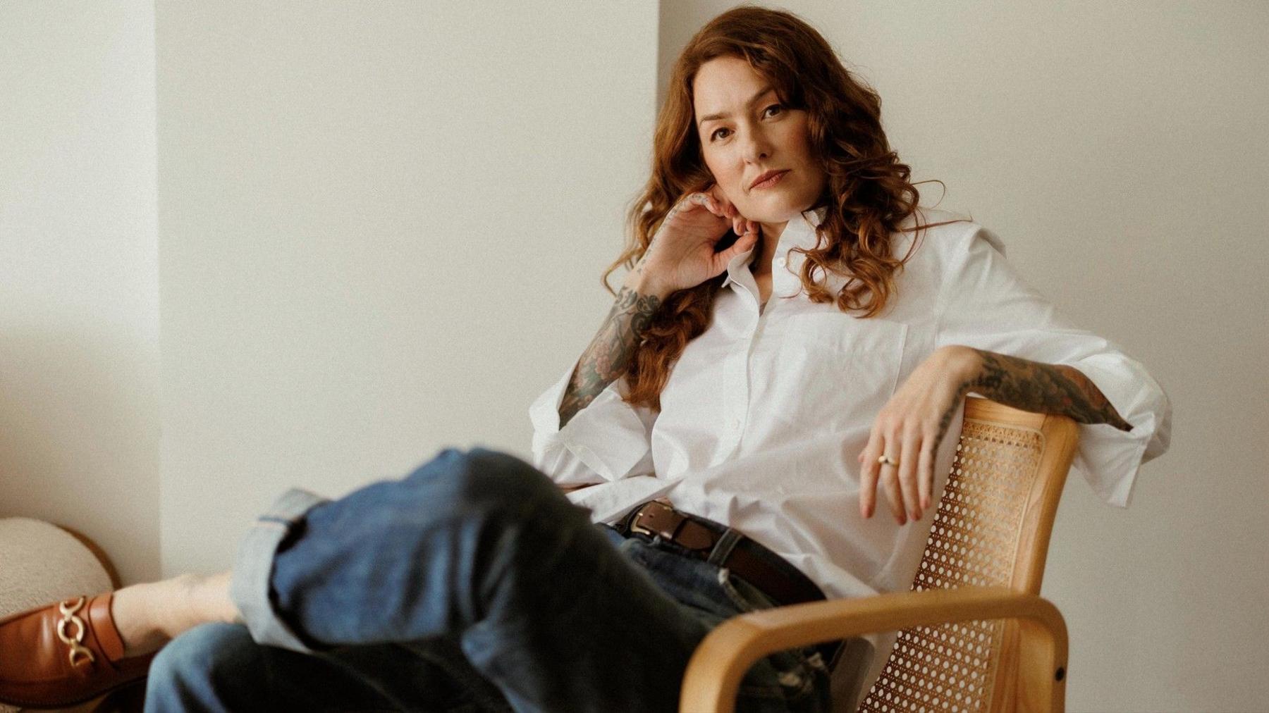 Maisie is sat on a wicker chair in a room painted white. She is wearing a white shirt with jeans and brown shoes. She has long red hair and tattooed arms.