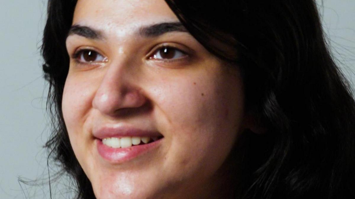 Iqra smiles and looks off camera. Her skin is clear. She has black hair and brown eyes.