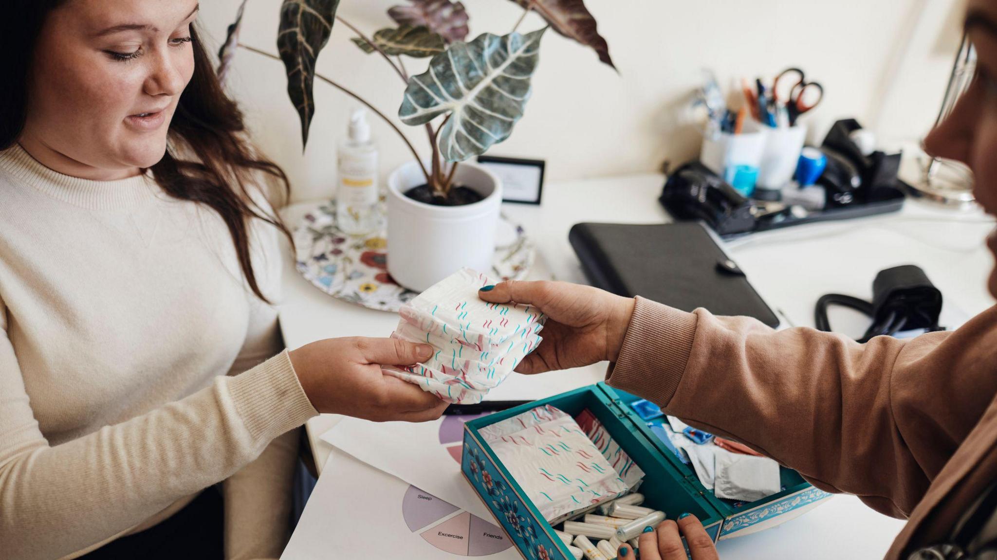 A woman, who is slightly off camera to the right, handing another woman who is in focus to the left, sanitary pads. There is a box of sanitary products in a box on a table in front of him. The woman on the left is wearing a white top and has long dark hair.