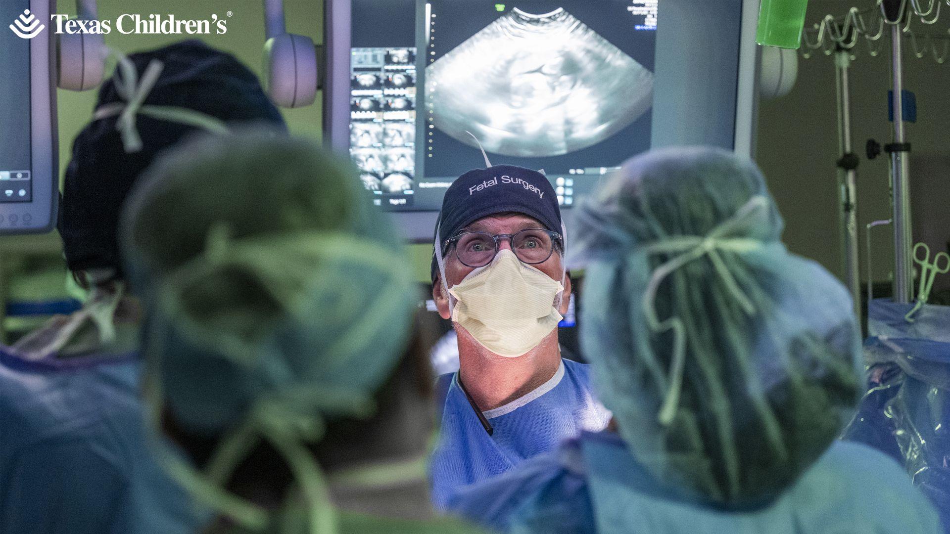 Medical staff in the foreground wearing surgical gowns, face masks and hair covers, gathered around an operating table. A person at the centre wears glasses, a face mask and a surgical cap labelled “Fetal Surgery”. Behind the team, large medical monitors display ultrasound images and diagnostic data. IV poles with hanging fluid bags and surgical instruments are visible at the sides of the operating theatre. The text “Texas Children’s” appears in the top-left corner of the image.