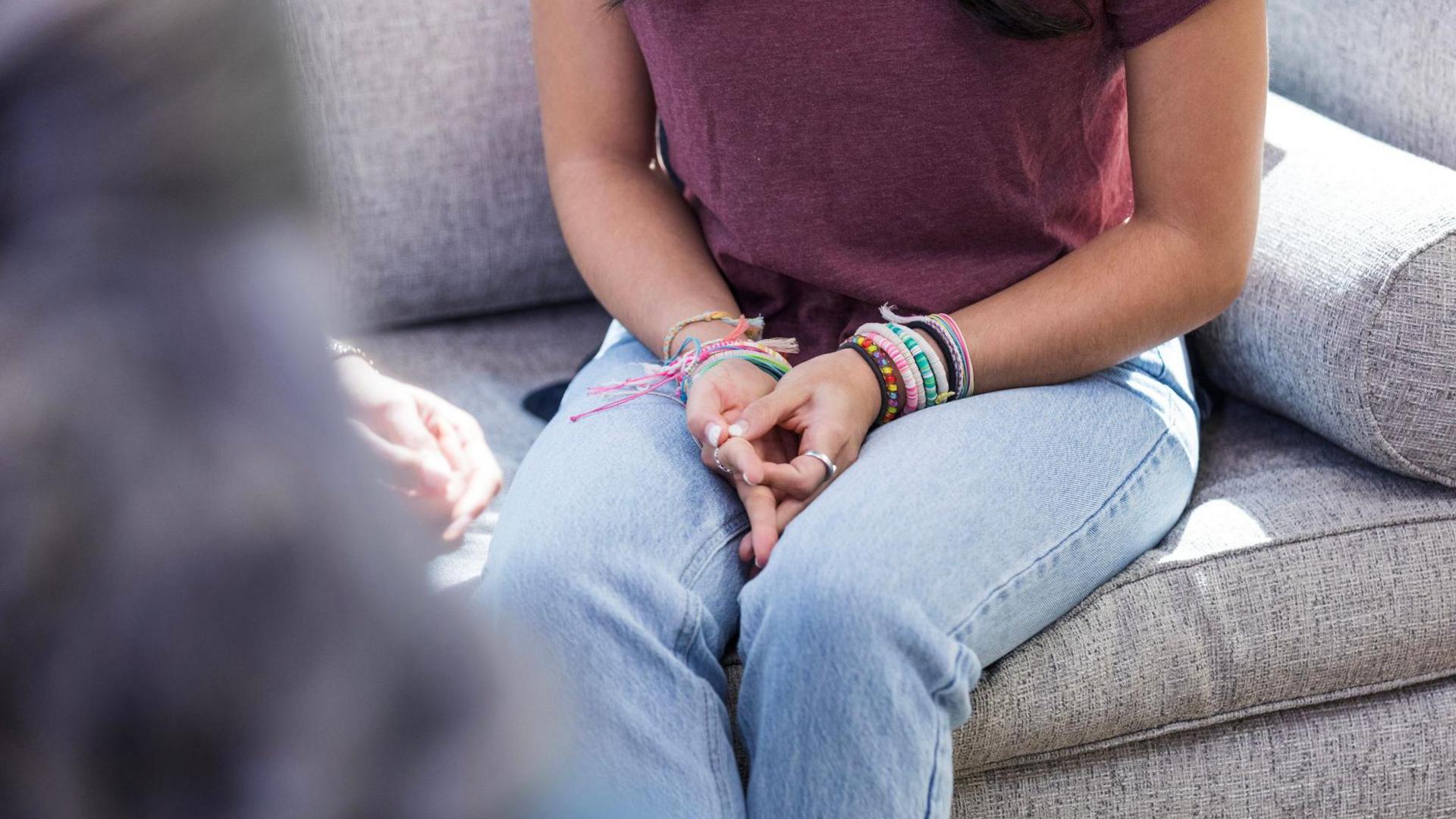A young woman with long dark hair in a burgundy t-shirt and light blue jeans sits on a grey sofa and claps her hands.