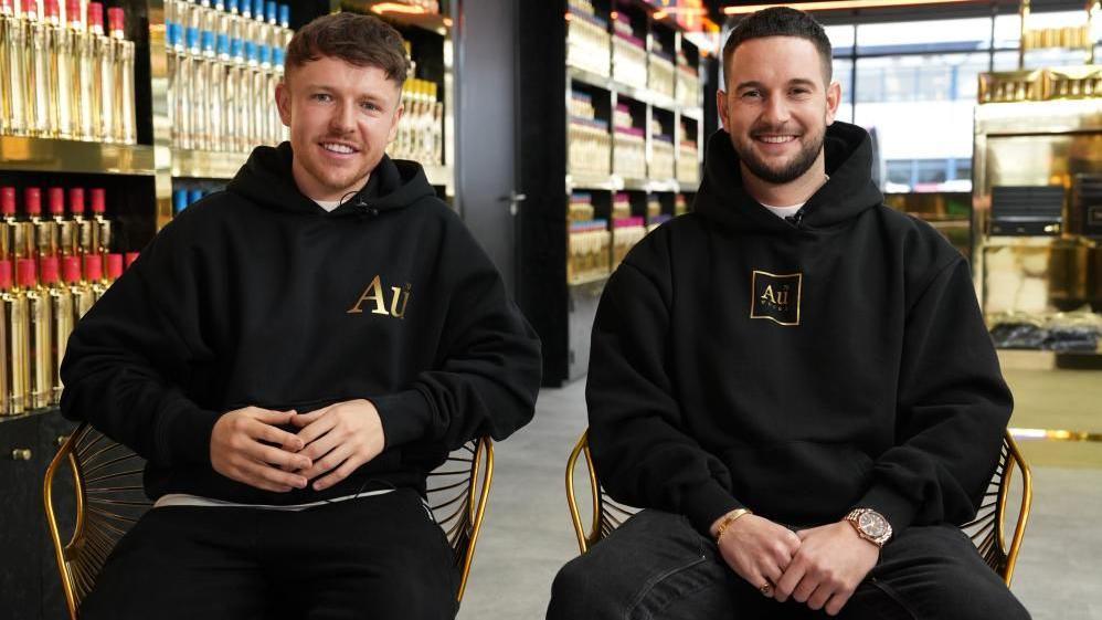 Two young men are smiling as they sit in golden framed chairs next to a row of gold-coloured Au Vodka bottles. They are both wearing black hoodies with the gold AU Vodka logo on them and black trousers.