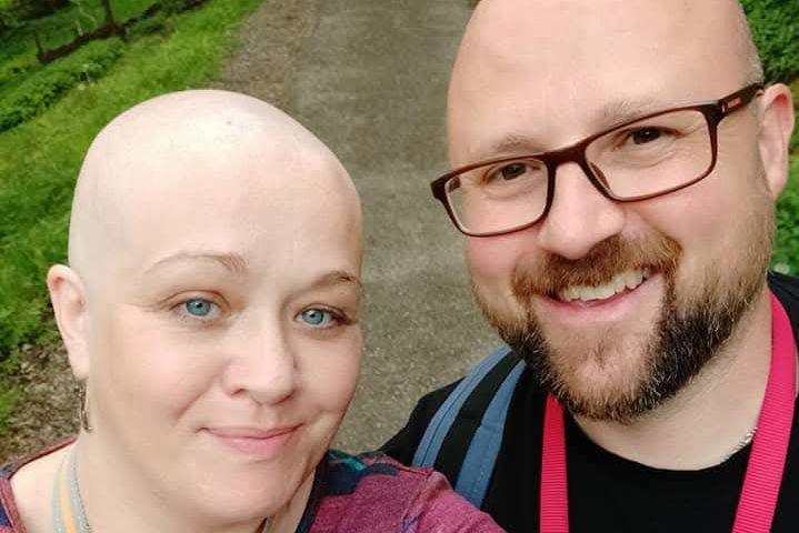 A woman with no hair is standing next to a man with a brown beard. They both appear to be standing in a park.