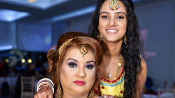 A woman with long dark hair, jewellery, and a yellow dress with her arm on the shoulder of a woman with ginger hair and jewellery