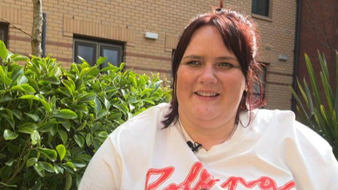 A woman with dark brown hair to her shoulders, in a white t-shirt with pink lettering, sitting in a garden next to a hedge. Brown-brick flats can be seen behind her. The woman is smiling at someone in front of her to her right and wearing a small microphone on the collar of her t-shirt.