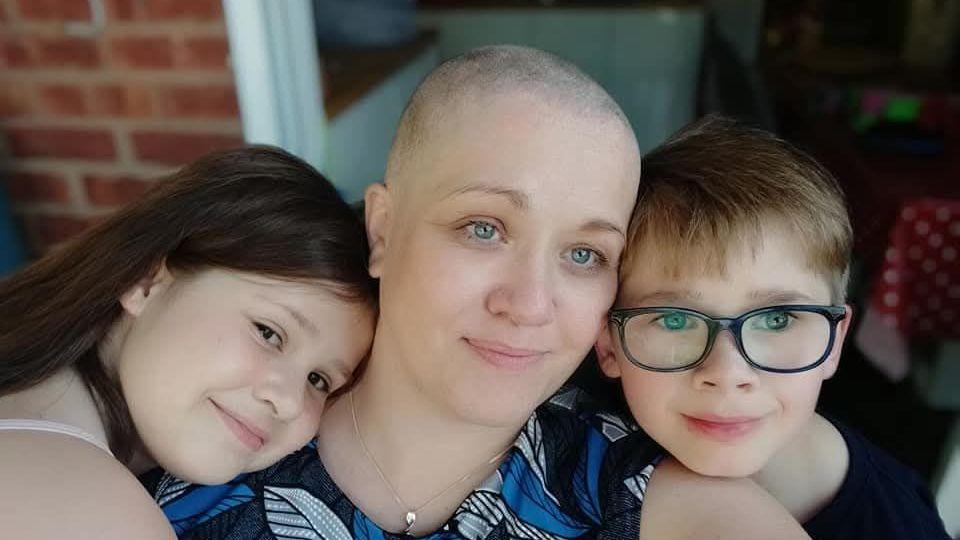 A woman who has lost her hair is being hugged by two children. They appear to be standing outside a house.