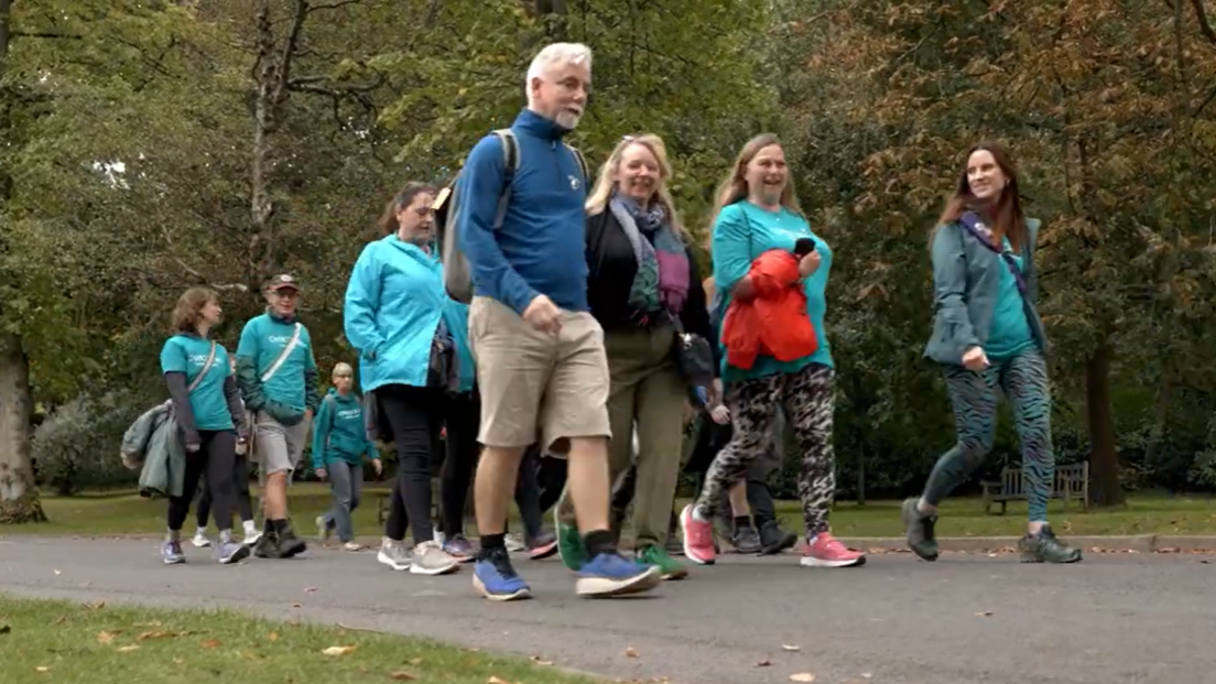 A group of people, mostly in teal T-shirts, are walking in a group along a path in a park.