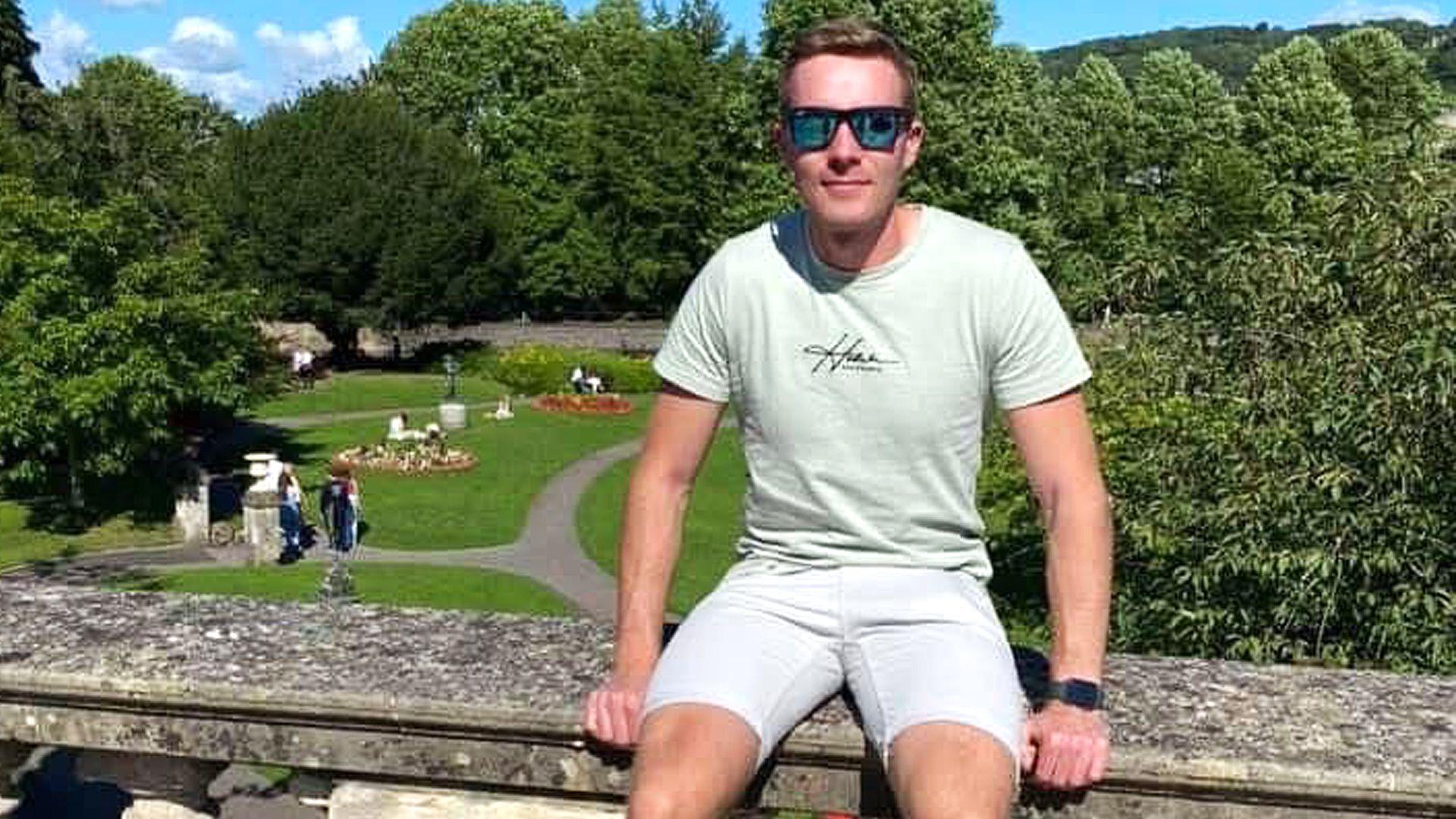 A young man sits on a stone balustrade with landscaped grounds behind him. He wears sunglasses, pale green T-shirt and white shorts. 