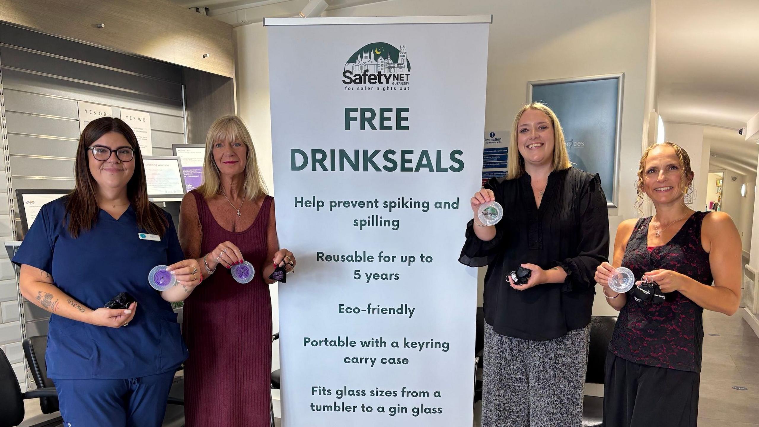 Four women stood next to a banner which says "Free Drinkseals" on it, holding up the covers that prevent drink spiking. They are wearing smart-casual clothing and two of the women are brunette whilst two of them are blonde.