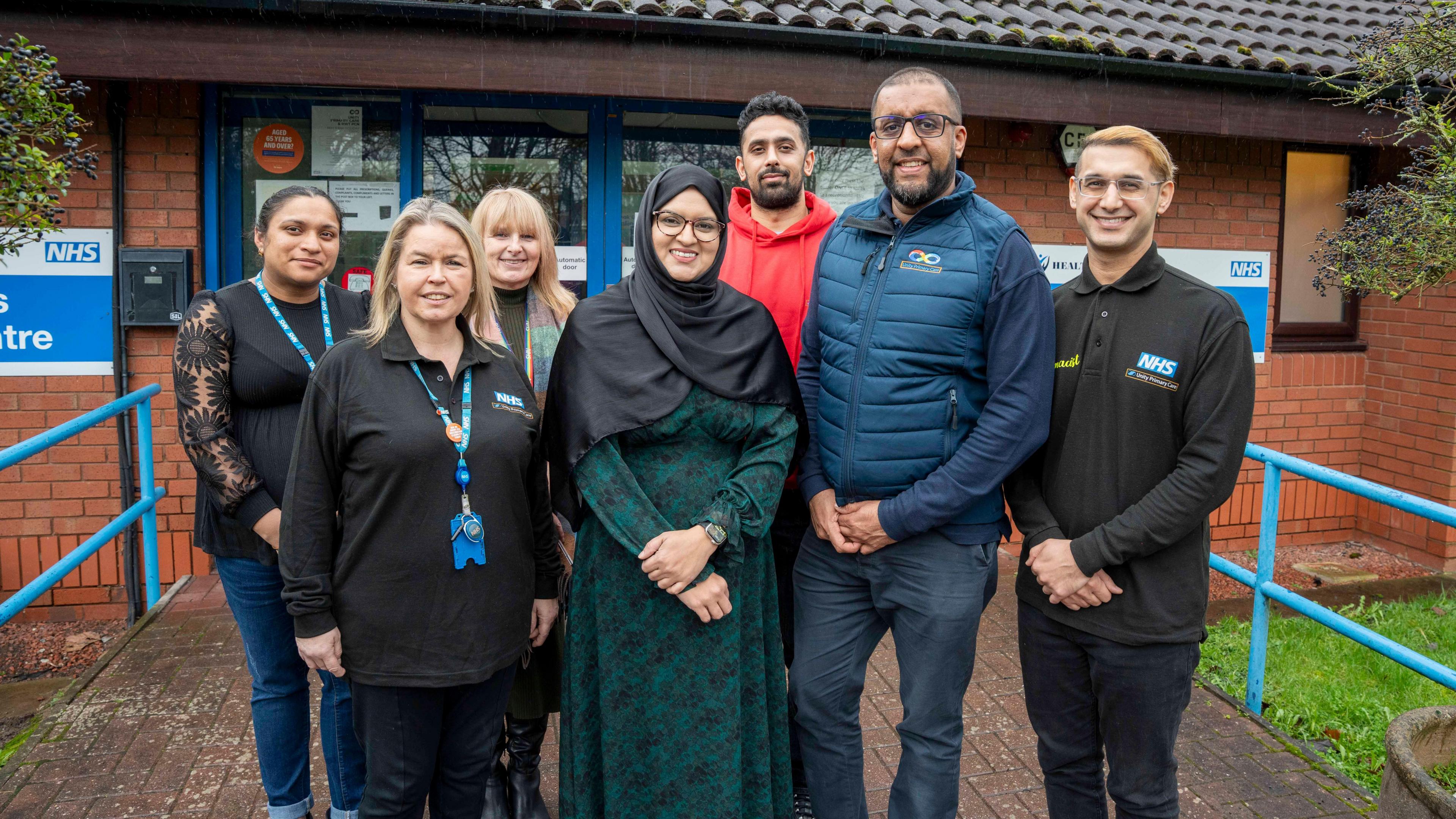 A group of seven people stand together outside an NHS health centre, several wearing NHS-branded clothing and lanyards. It is a bright day.