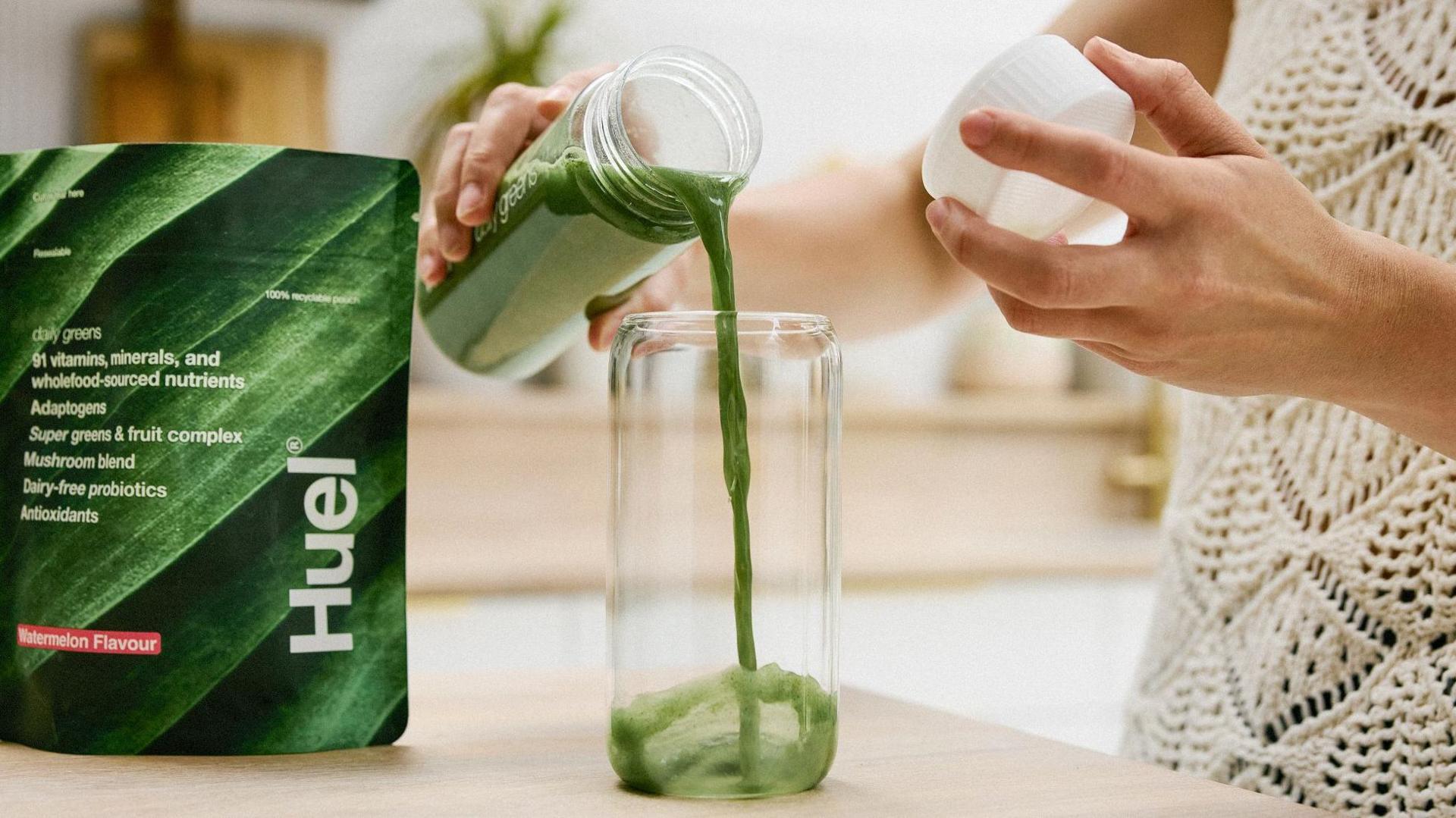 A green nutritional drink is being poured into a glass. The setting appears to be a bright, modern kitchen or dining area with a blurred background.