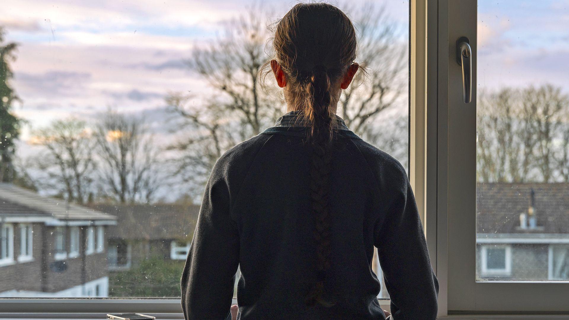 A young girl stands at a window and looks out on to a neighbourhood
