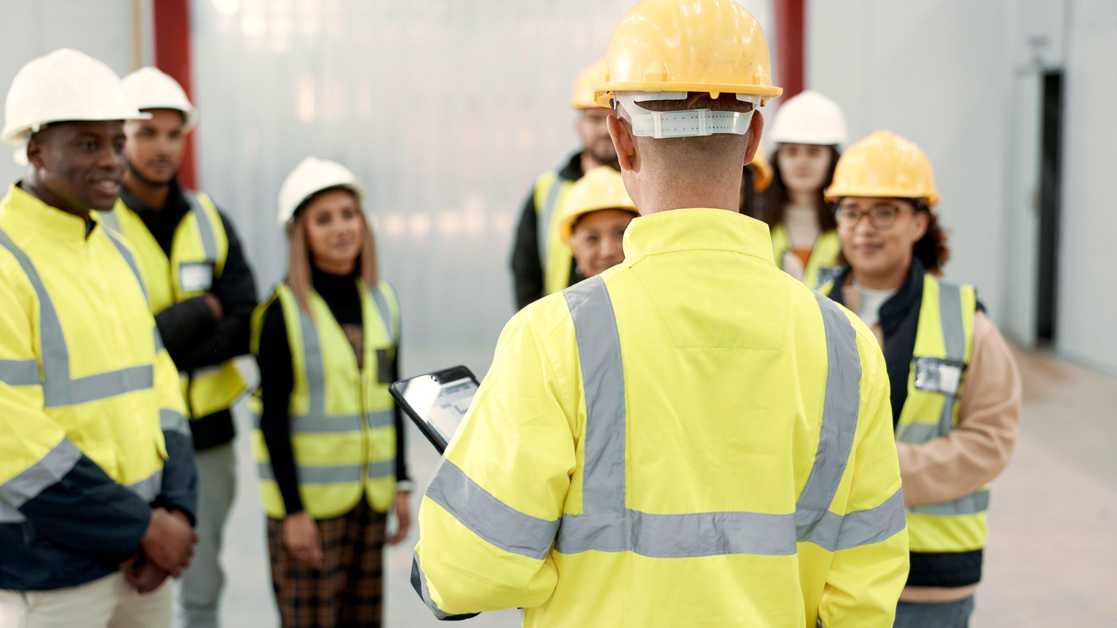 A group of people wearing hi-vis yellow jackets stand around in a room. Most of the group are standing facing a man with a clipboard, whose back is towards the camera. They are listening to what he is saying.
