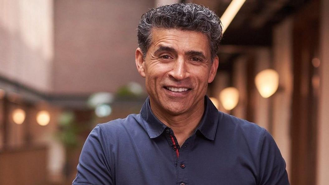 Professor Ama Johal, a man with dark, grey hair which is cut short, smiles at the camera. He is wearing a navy collared polo shirt and the background is blurred.