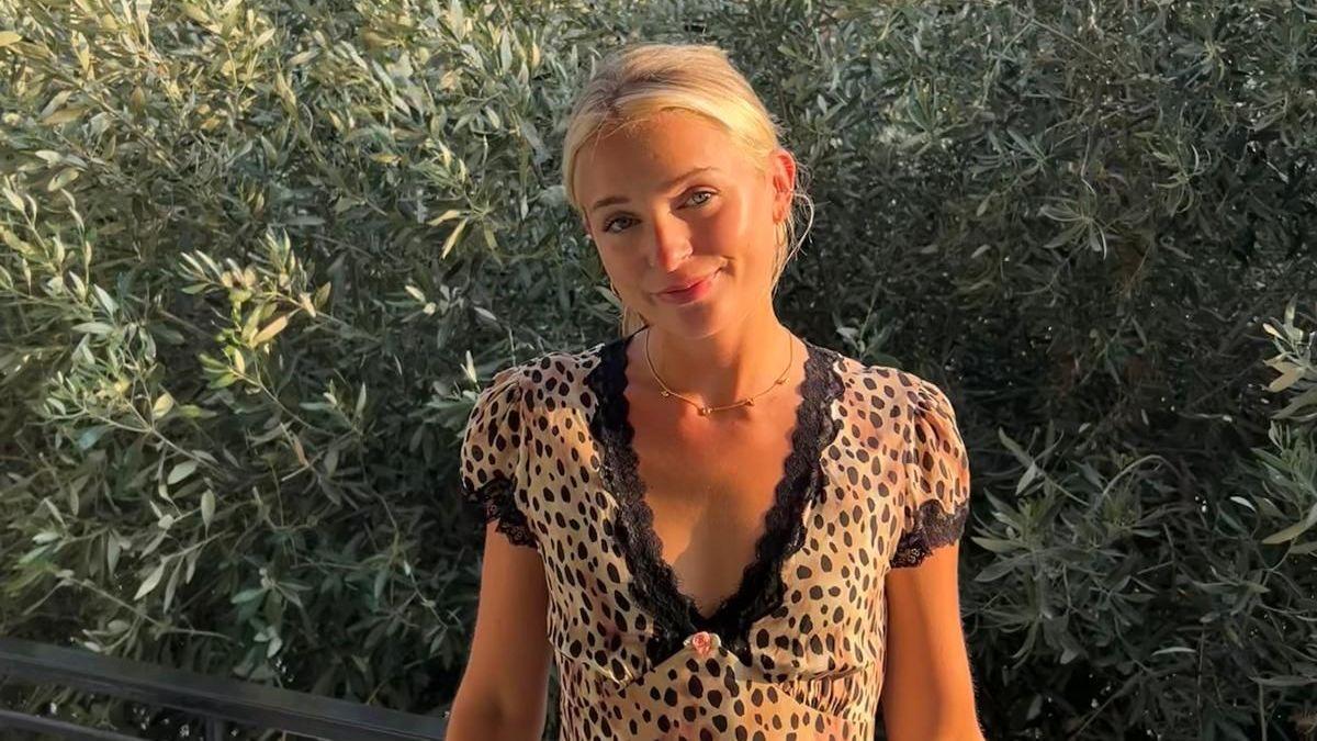 Meg Warren, a young woman with blonde hair, is standing in front of a bush and smiling. She is wearing a leopard print dress.