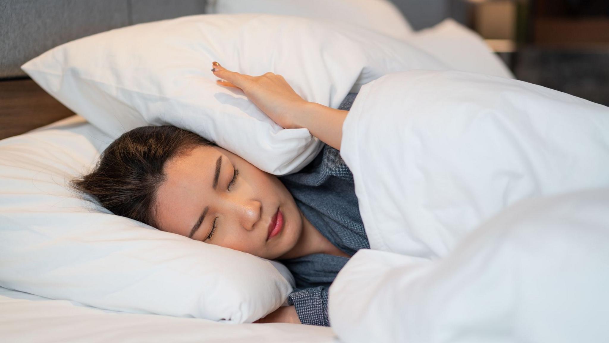 A woman with dark hair lies in bed with her head on a white pillow and a white duvet covering her body. Her eyes are closed and she is lying on her side while holding another white pillow over one side of her head as if to block out noise.